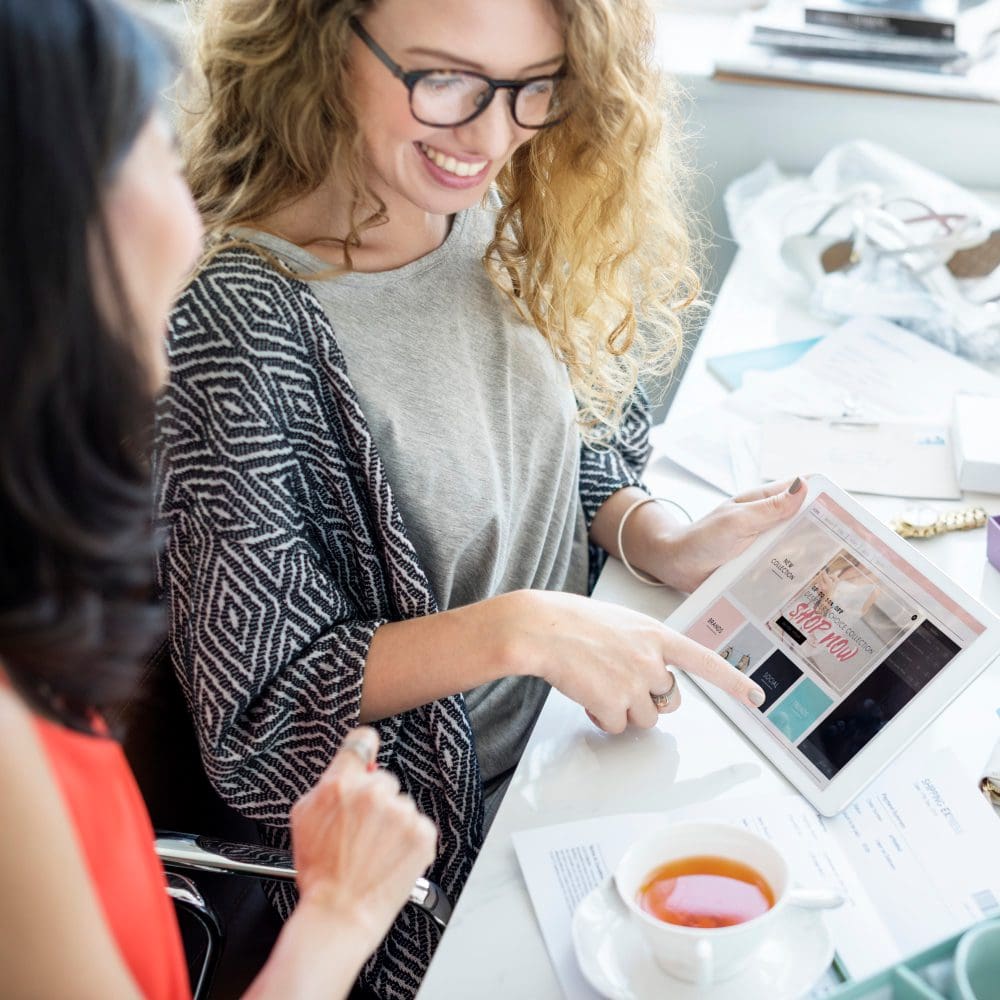 Two women engaged in local digital marketing study while examining a tablet at a desk. Two women engaged in local digital marketing study while examining a tablet at a desk.