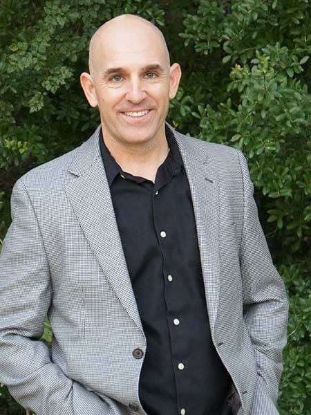Brian Gibbs is smiling while posing with his hands in his pockets. He is wearing a black shirt and a gray plaid blazer, embodying the essence of smart business style. Behind him, the dense green foliage adds a refreshing touch to this Smart Business Accelerator promotional shoot. Brian Gibbs is smiling while posing with his hands in his pockets. He is wearing a black shirt and a gray plaid blazer, embodying the essence of smart business style. Behind him, the dense green foliage adds a refreshing touch to this Smart Business Accelerator promotional shoot.