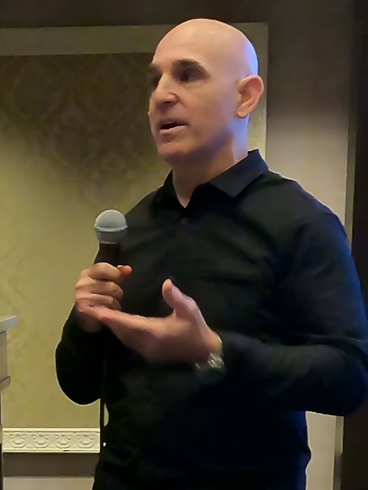 A bald man in a dark shirt holds a microphone and speaks at an event. Digital marketing consultant Brian Gibbs is gesturing with his other hand, and a beige wall with decorative molding is visible in the background. A bald man in a dark shirt holds a microphone and speaks at an event. Digital marketing consultant Brian Gibbs is gesturing with his other hand, and a beige wall with decorative molding is visible in the background.
