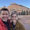 A couple smiling and taking a selfie with a scenic desert landscape featuring a large red rock formation and a clear blue sky in the background.